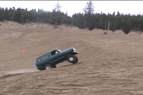 1979 Ford Bronco - A green Ford Bronco is shown navigating a steep sand dune, kicking up dust as it ascends.