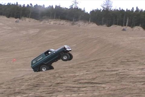 1979 Ford Bronco - A green SUV is airborne on a sand dune, with another vehicle visible in the background.