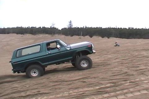 1979 Ford Bronco - A lifted green Ford Bronco navigates sandy terrain under a cloudy sky.