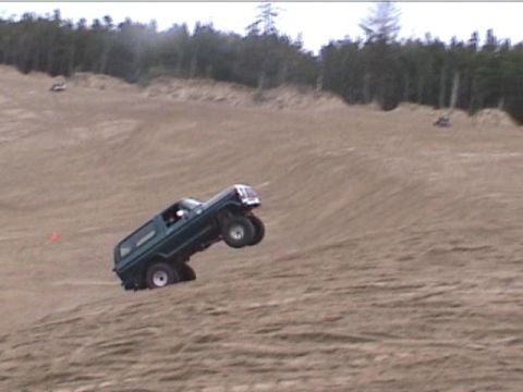 1979 Ford Bronco - A dark green SUV navigates a steep sandy incline, with its front wheels airborne.