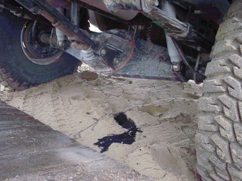 1979 Ford Bronco - Vehicle undercarriage stuck in sand, leaking dark fluid. Tire tracks visible in the sand.