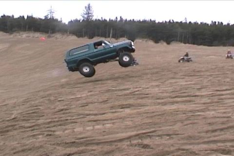 1979 Ford Bronco - A green Ford Bronco is captured mid-air as it jumps over a sand dune, with ATVs visible in the distance.
