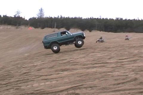 1979 Ford Bronco - A green SUV with lifted suspension is airborne on a sand dune, with two ATVs visible in the background.