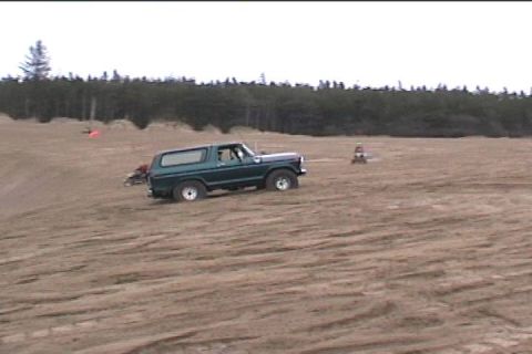 1979 Ford Bronco - A green 1979 Ford Bronco navigates a sandy landscape, with trees in the background.