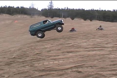 1979 Ford Bronco - Ford Bronco airborne over a sand dune, with ATVs in the distance.