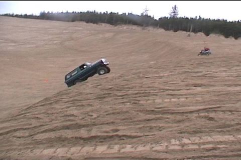 1979 Ford Bronco - A dark-colored SUV is captured mid-air as it ascends a sandy incline, with an ATV visible in the distance.
