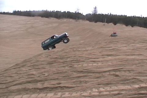 1979 Ford Bronco - A dark green SUV is airborne while cresting a sand dune, with another vehicle visible in the background.