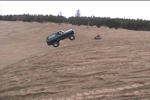 1979 Ford Bronco - SUV airborne during a jump, with an ATV visible in the distance on a sandy terrain.