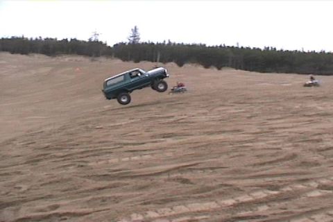 1979 Ford Bronco - An off-road vehicle catches air while traversing a sandy terrain, with other recreational vehicles visible in the distance.