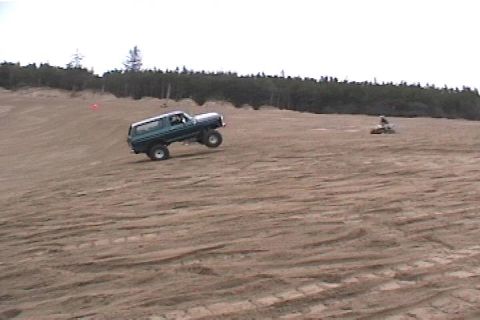 1979 Ford Bronco - A green SUV navigates a sandy incline with a wooded area in the background and a distant ATV rider.