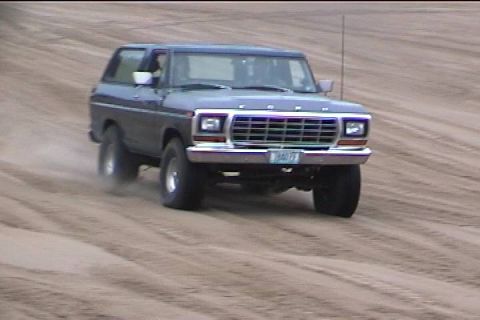 1979 Ford Bronco - A blue Ford Bronco drives on a sandy terrain, with dust trailing behind it.