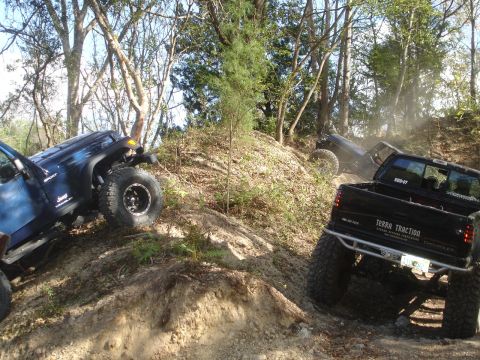 1989 Ford Bronco - Several off-road vehicles navigate a steep, dusty incline surrounded by trees.