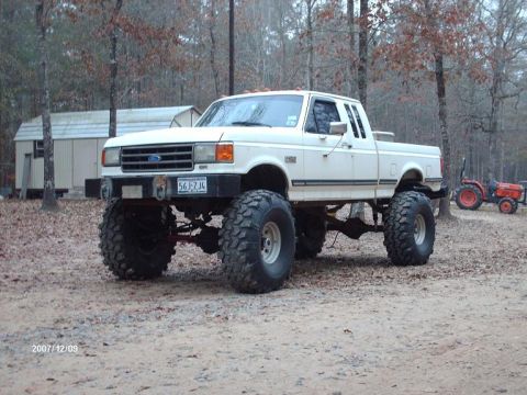 1990 Ford F-150 - A lifted white pickup truck with oversized tires sits in a wooded area next to two tractors.