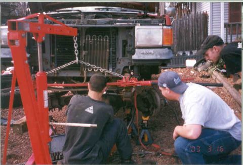 1991 Ford F-250 - Two men are working on the front suspension of a truck, using a red engine hoist.