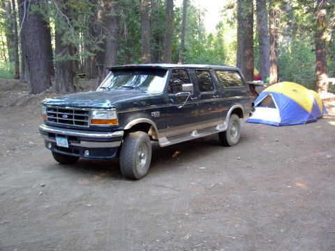 1995 Ford C-350 (Centurion Conversions) - A dark blue 1995 Ford C-350 Centurion Conversions SUV is parked at a campsite next to a blue and yellow tent.