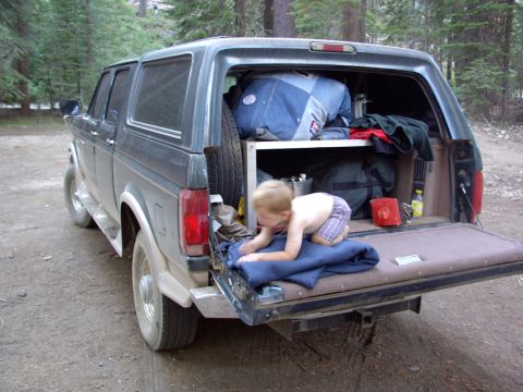 1995 Ford C-350 (Centurion Conversions) - A young child plays on a blanket in the open tailgate of a green pickup truck packed for a camping trip.