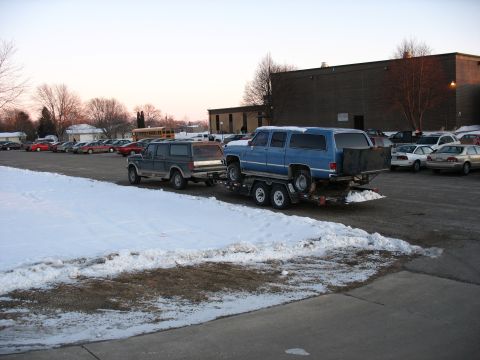 1995 Ford C-350 (Centurion Conversions) - A dark gray pickup truck tows a blue SUV on a trailer in a snowy parking lot.