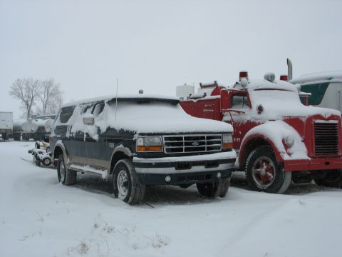 1995 Ford C-350 (Centurion Conversions) - A snow-covered Ford Bronco sits parked next to a vintage red fire truck in a winter scene.