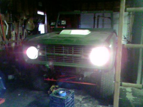 1974 Ford Bronco - A green SUV with a distinctive grille and hood scoop is parked indoors, its headlights illuminated.