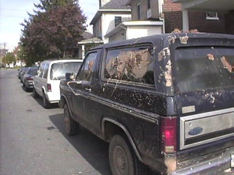 1985 Ford Bronco - A black Ford Bronco with significant body damage and mud-caked windows is parked on a residential street.