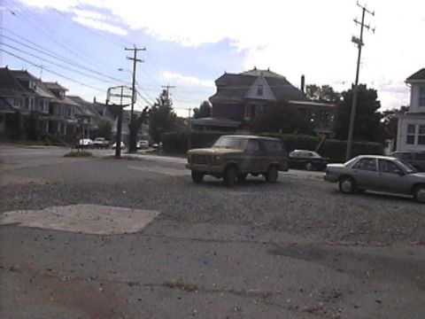 1985 Ford Bronco - A brown Ford Bronco is parked on a gravel lot, with houses and other vehicles visible.