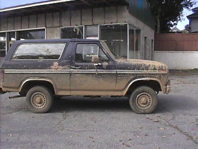 Mud-covered dark blue Ford Bronco, side view, showing rust spots and flat front tire