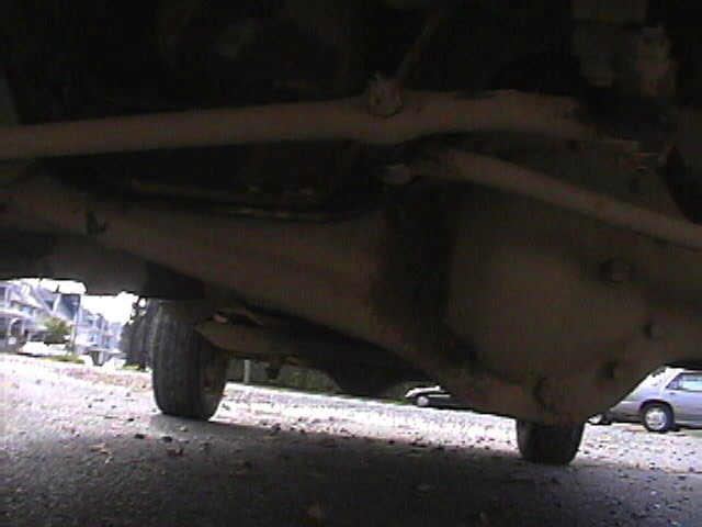Underside view of a vehicle's drivetrain and suspension components. Another car is visible in the background