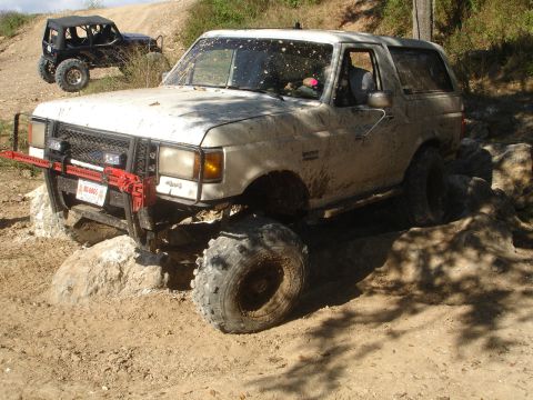 1989 Ford Bronco - A muddy 1989 Ford Bronco tackles a rocky off-road course, with a second vehicle visible in the background.