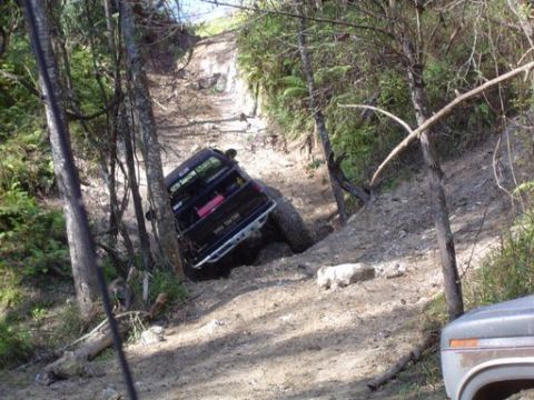 1989 Ford Bronco - A black off-road vehicle is shown struggling on a steep, muddy incline surrounded by trees.