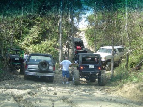 1989 Ford Bronco - Several off-road vehicles navigate a muddy trail through a wooded area, with a 1989 Ford Bronco visible.