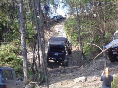 1989 Ford Bronco - Multiple off-road vehicles are seen on a challenging, muddy trail surrounded by dense trees.