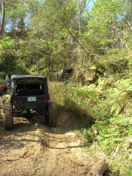 1989 Ford Bronco - An off-road vehicle navigates a muddy trail surrounded by dense foliage, with another vehicle visible in the distance.