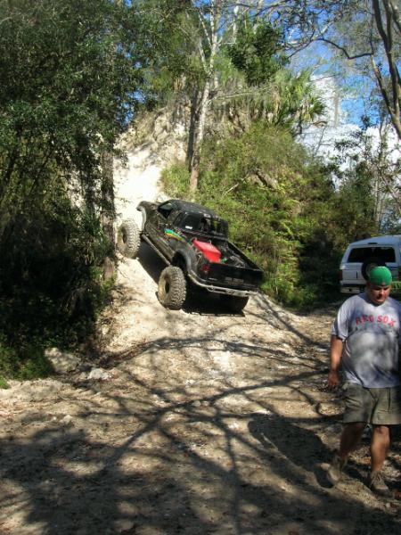 1989 Ford Bronco - A black off-road truck navigates a challenging incline on a dirt trail, with another vehicle and a person visible in the background.