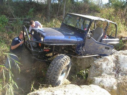 1989 Ford Bronco - Two people attempt to free a blue Jeep Wrangler that is stuck on a rocky incline during an off-road excursion.