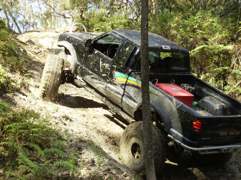 Black pickup truck with large tires climbing a steep, muddy, wooded trail. It has a red fuel tank in the bed