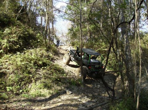 1989 Ford Bronco - An off-road vehicle navigates a challenging, muddy incline through a wooded area.