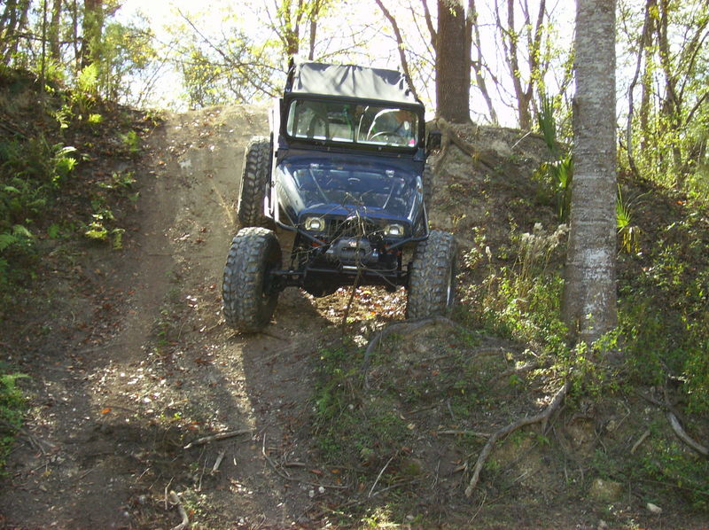 Blue Jeep with large tires climbing a dirt hill, soft top up, front view