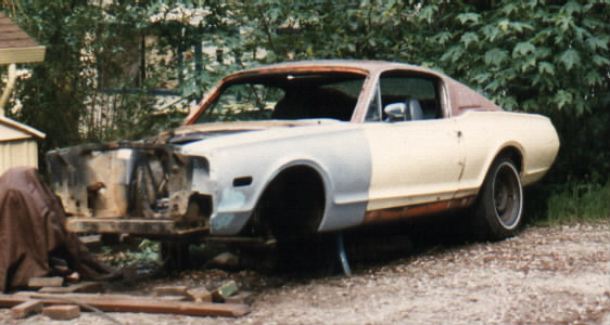 Partially disassembled white 1968 Mercury Cougar project car with primer and rust, missing front end parts, on gravel