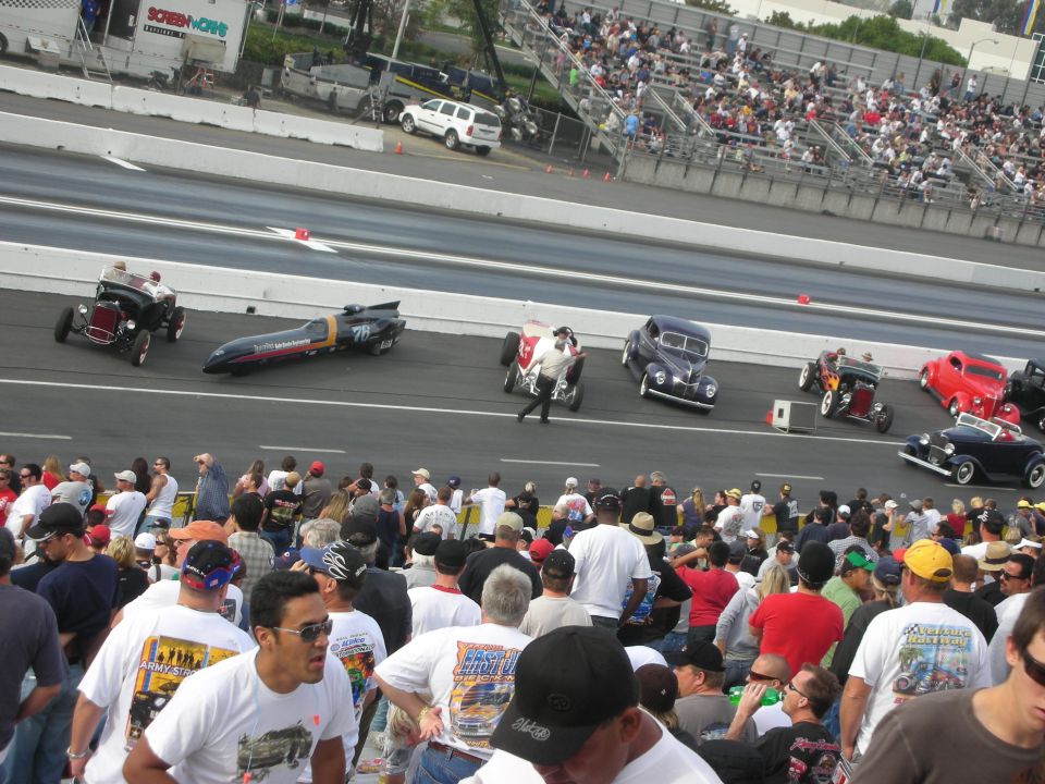 Vintage hot rods and a streamliner lined up on a drag strip before a crowd