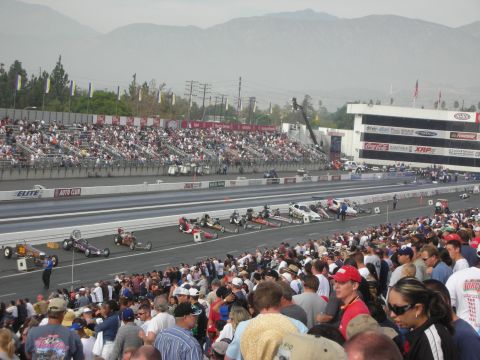 1998 Ford Crown Victoria - Front-engine dragsters stage on a drag strip with packed grandstands and mountains in the background.