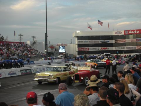 1998 Ford Crown Victoria - yellow gasser and a red hot rod roadster sit at the starting line of a drag strip as spectators watch at dusk.