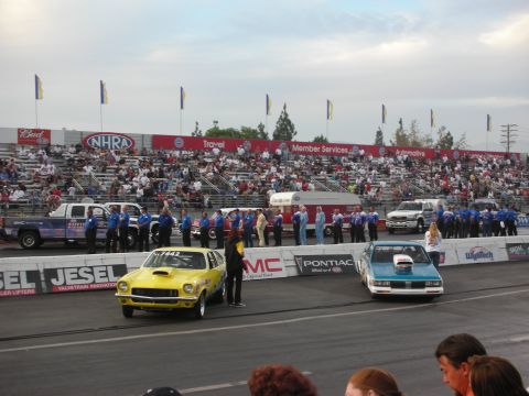 1998 Ford Crown Victoria - yellow and a teal vintage drag car stage at the starting line as officials in blue shirts line the track.