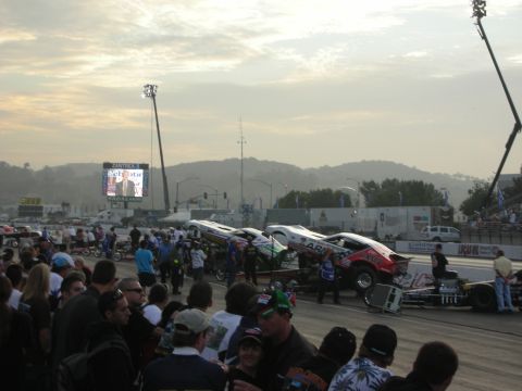 1998 Ford Crown Victoria - row of funny cars sits staged along a drag strip at dusk while a crowd of spectators watches.