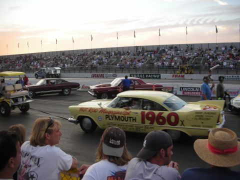 1998 Ford Crown Victoria - yellow Plymouth gasser lettered "Hot Rod Magazine 1960" stages at the drag strip line at sunset before a crowd.