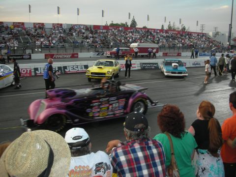 1998 Ford Crown Victoria - purple hot rod roadster launches down the drag strip as yellow and teal gassers stage behind, spectators watching.