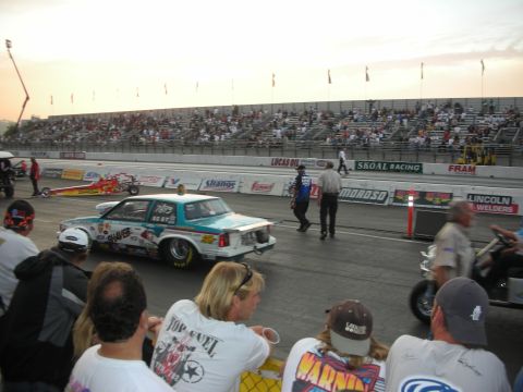 1998 Ford Crown Victoria - teal door-slammer drag car and a front-engine dragster stage at the line as spectators watch at dusk.