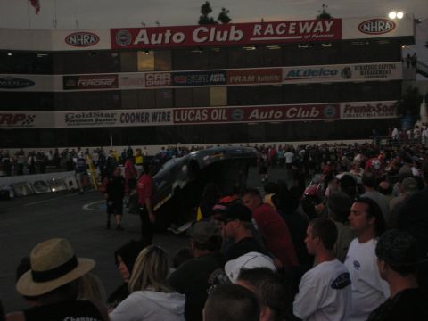 1998 Ford Crown Victoria - Spectators crowd the fence watching a funny car at Auto Club Raceway at Pomona under sponsor banners at dusk.