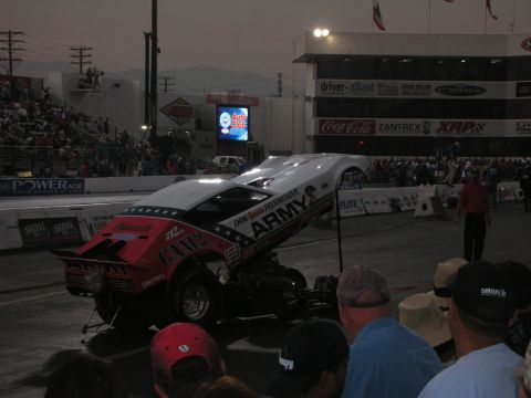1998 Ford Crown Victoria - red ARMY-sponsored funny car lifts its front wheels off the line on the drag strip as spectators watch at dusk.