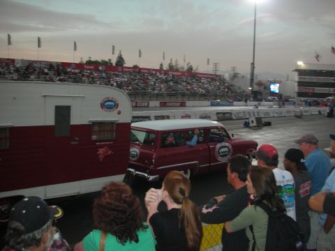 1998 Ford Crown Victoria - maroon vintage station wagon tows its matching trailer past spectators at the drag strip in fading light.