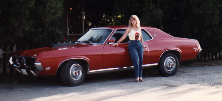 Red 1969 Mercury Cougar coupe parked on gravel with a woman leaning against the driver's side.